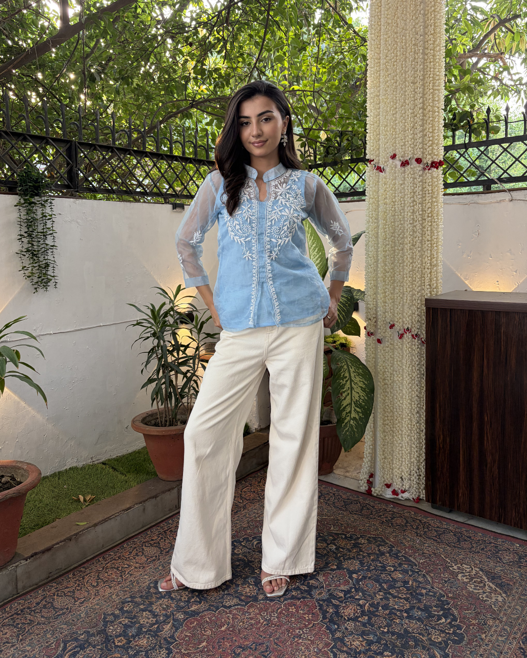 Woman in a light blue embroidered top and white pants standing in an outdoor setting with plants and a white wall.