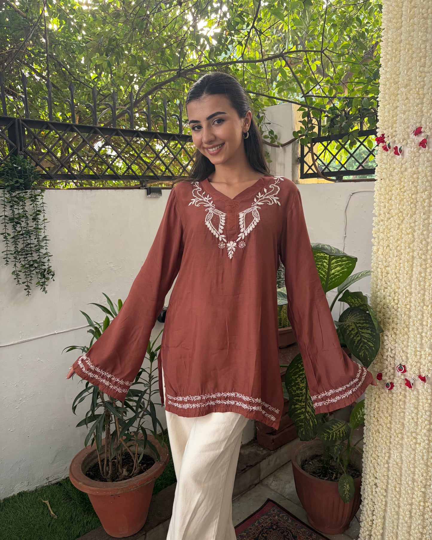 Woman wearing a brown embroidered top in an outdoor setting with plants and a white wall.