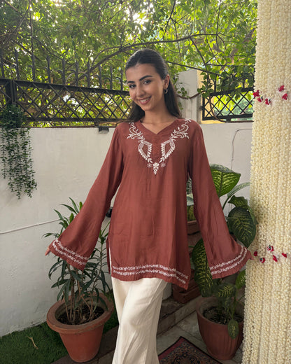 Woman wearing a brown embroidered top in an outdoor setting with plants and a white wall.