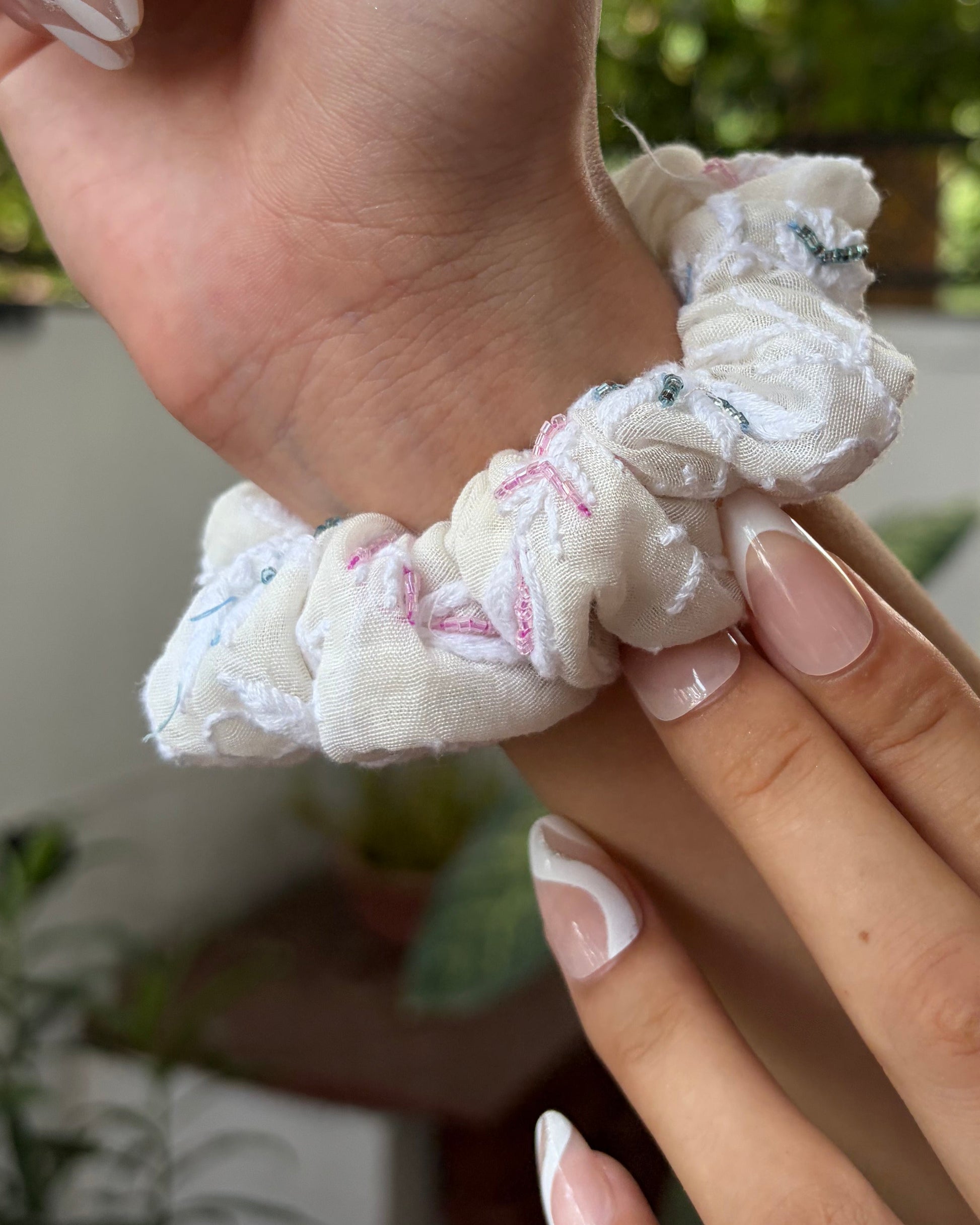 Hand wearing a white scrunchie with blurred greenery in the background