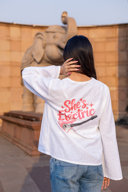 A woman viewed from behind wearing a white long-sleeve t-shirt with a pink graphic that reads 'She's Electric' and a guitar illustration, paired with blue jeans, standing in front of a stone elephant statue.