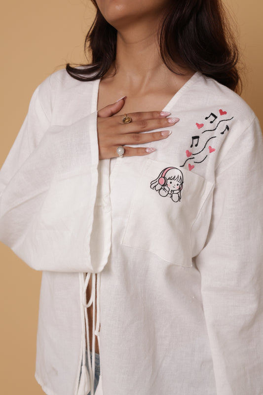 A close-up of a woman wearing a white linen open-front cardigan with delicate embroidery on the chest depicting a cartoon girl with headphones, musical notes, and pink hearts, set against a solid beige background.