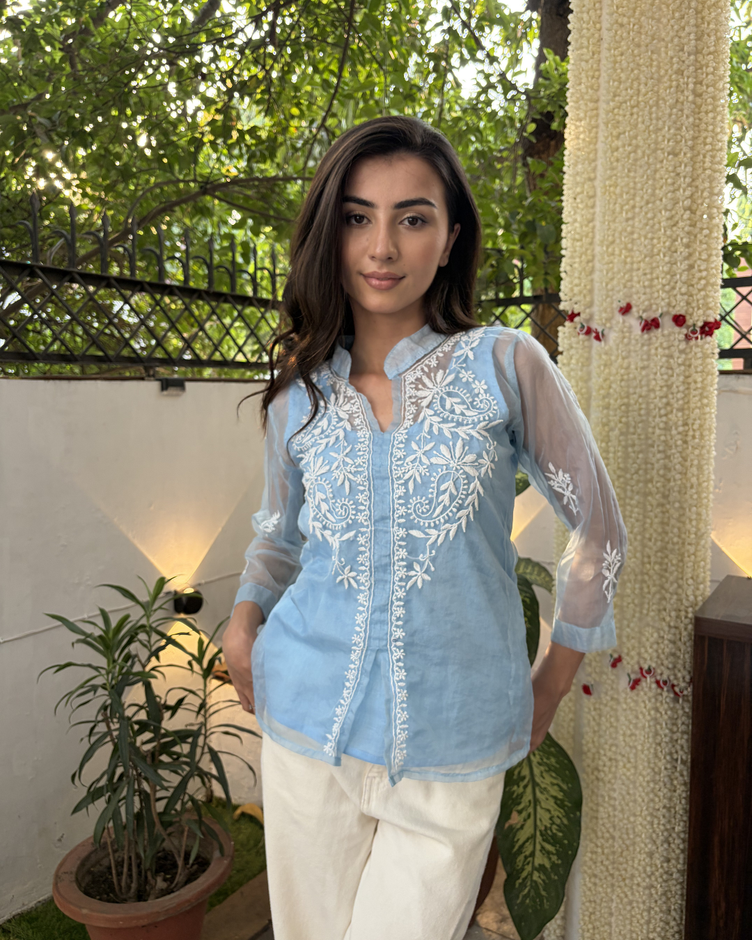 Woman wearing a light blue embroidered blouse with white pants standing outdoors.
