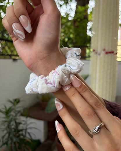 Hand wearing a white scrunchie with blurred greenery in the background